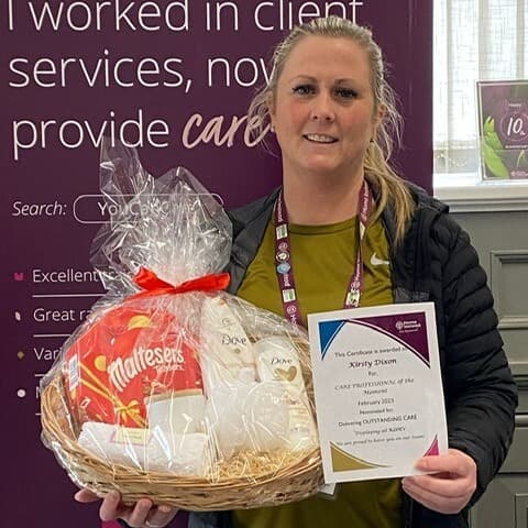 A woman holding a gift basket and a certificate in front of a display with text about client services. - Home Instead