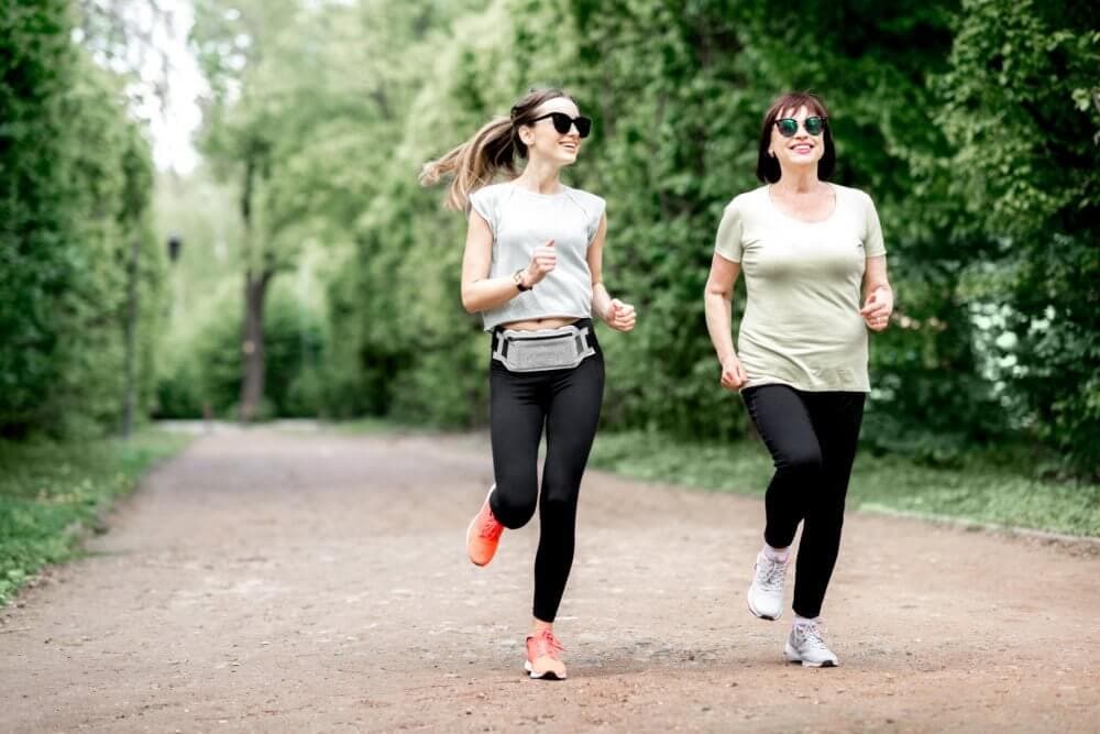 Two women in athletic wear jog along a tree-lined path on a sunny day, both smiling and wearing sunglasses. - Home Instead