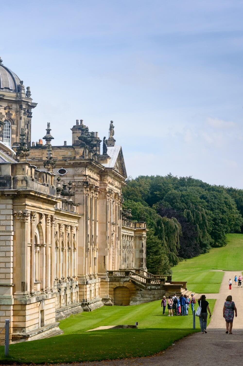 People walking near an ornate historic building with lush green lawns and a wooded area in the background. - Home Instead