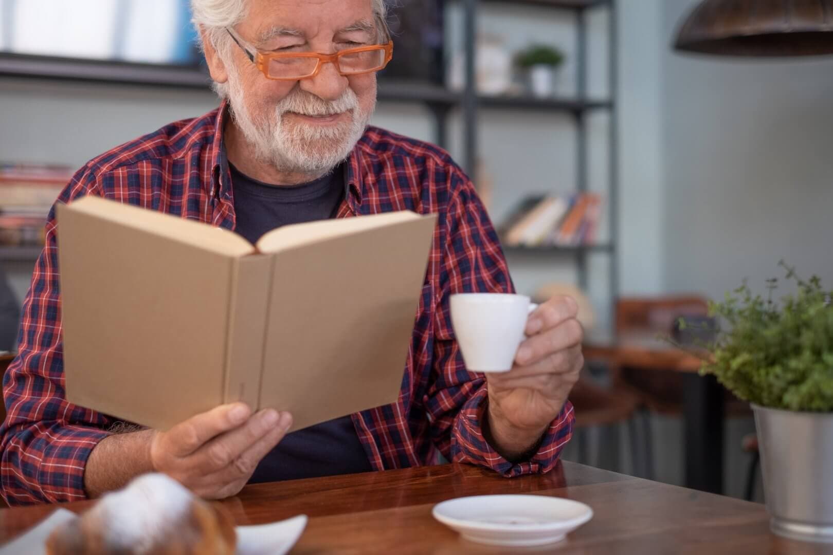 Smiling elderly man with glasses reading a book and holding a cup of coffee in a cozy, book-filled room. - Home Instead Bournemouth & Christchurch