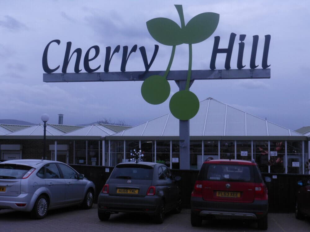 Large sign reading "Cherry Hill" with green cherry logo, above parked cars in front of a building. - Home Instead