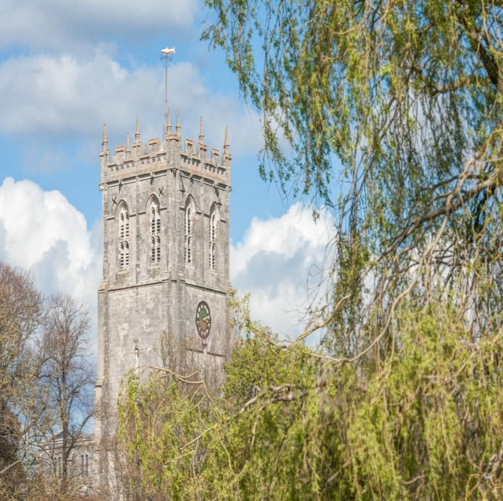 A stone clock tower with battlements stands tall behind trees, under a bright blue sky with fluffy clouds. - Home Instead