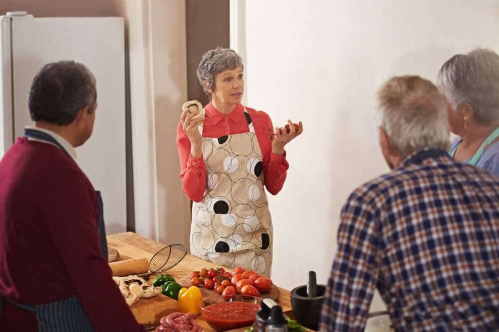 A woman in an apron instructs a cooking class, holding a bread roll, with three participants watching her. - Home Instead