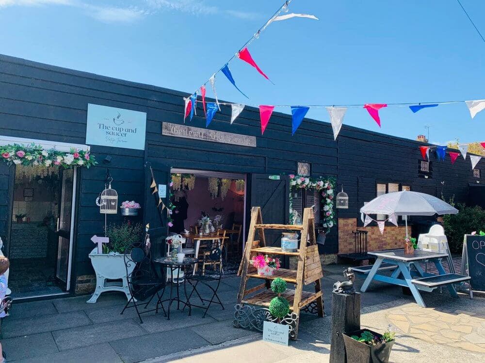 Outdoor view of a cozy café with patio seating, hanging flower pots, and colorful bunting on a sunny day. - Home Instead