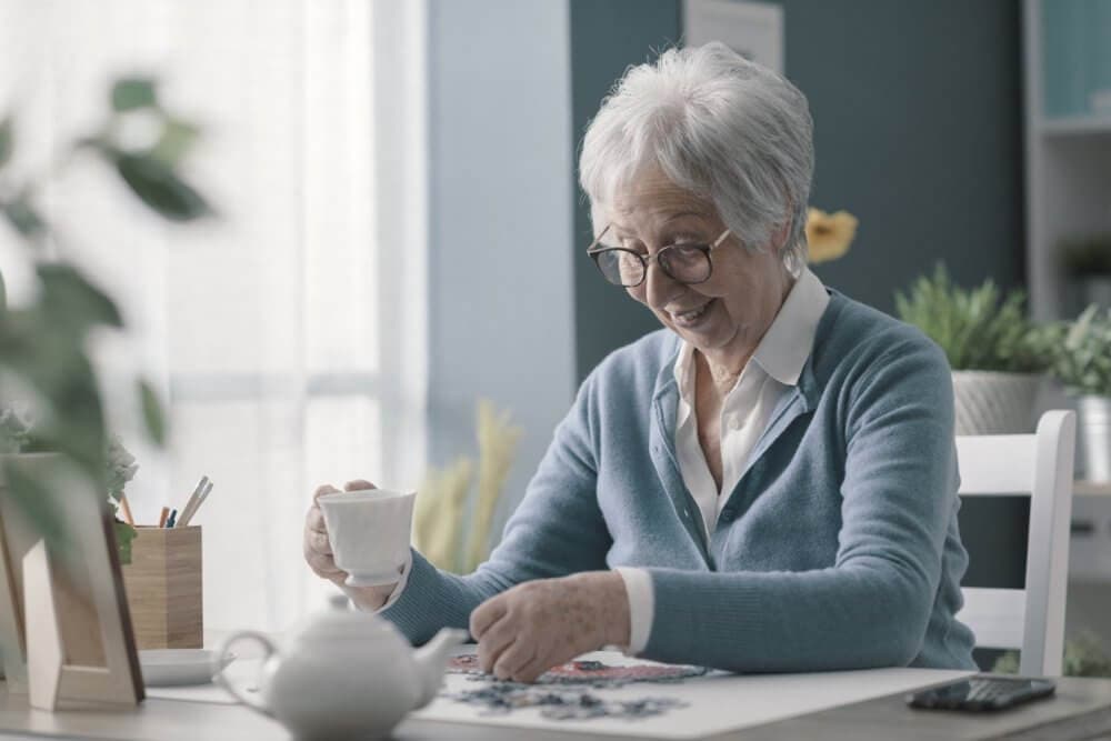 Elderly woman with glasses and a blue sweater drinks tea while working on a puzzle at a table. - Home Instead