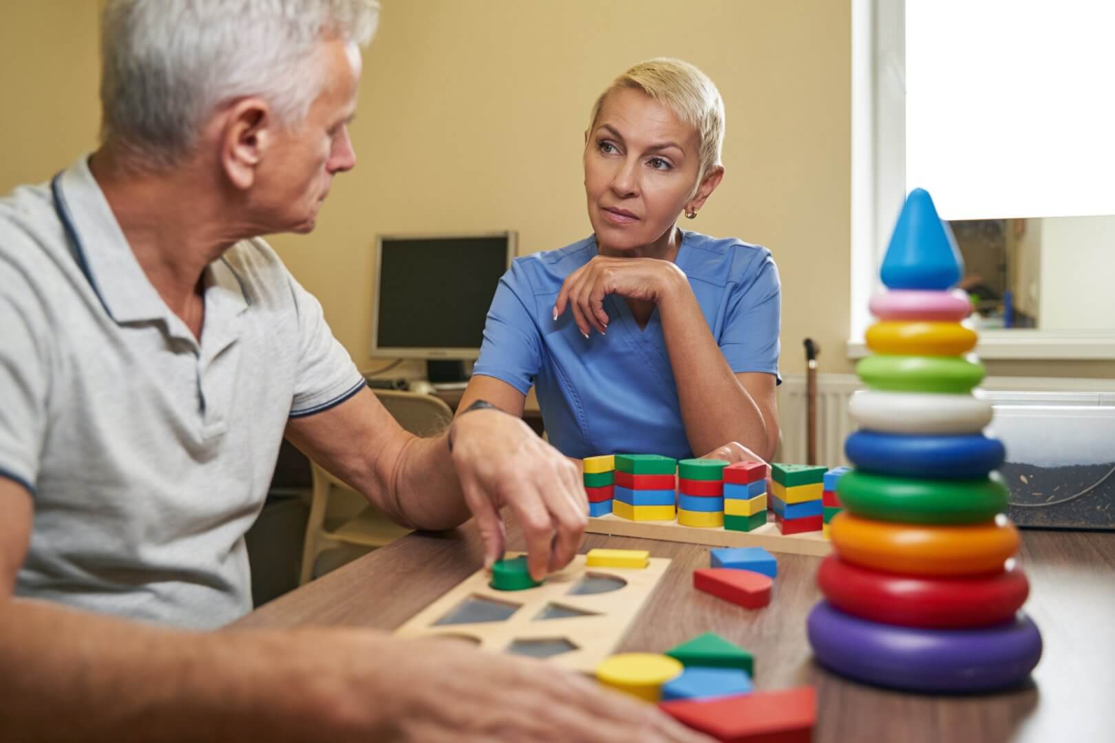 A man and a caregiver work on a puzzle at a table with various colorful geometric toys in a room. - Home Instead