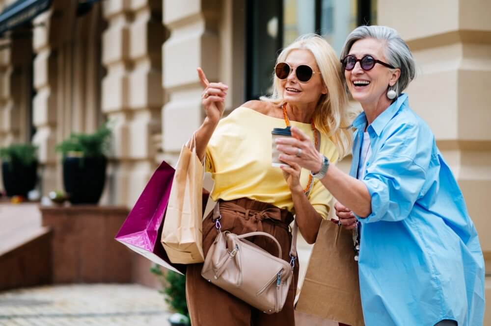 Two women with shopping bags and coffee cups, smiling and pointing while standing outside a building. - Home Instead