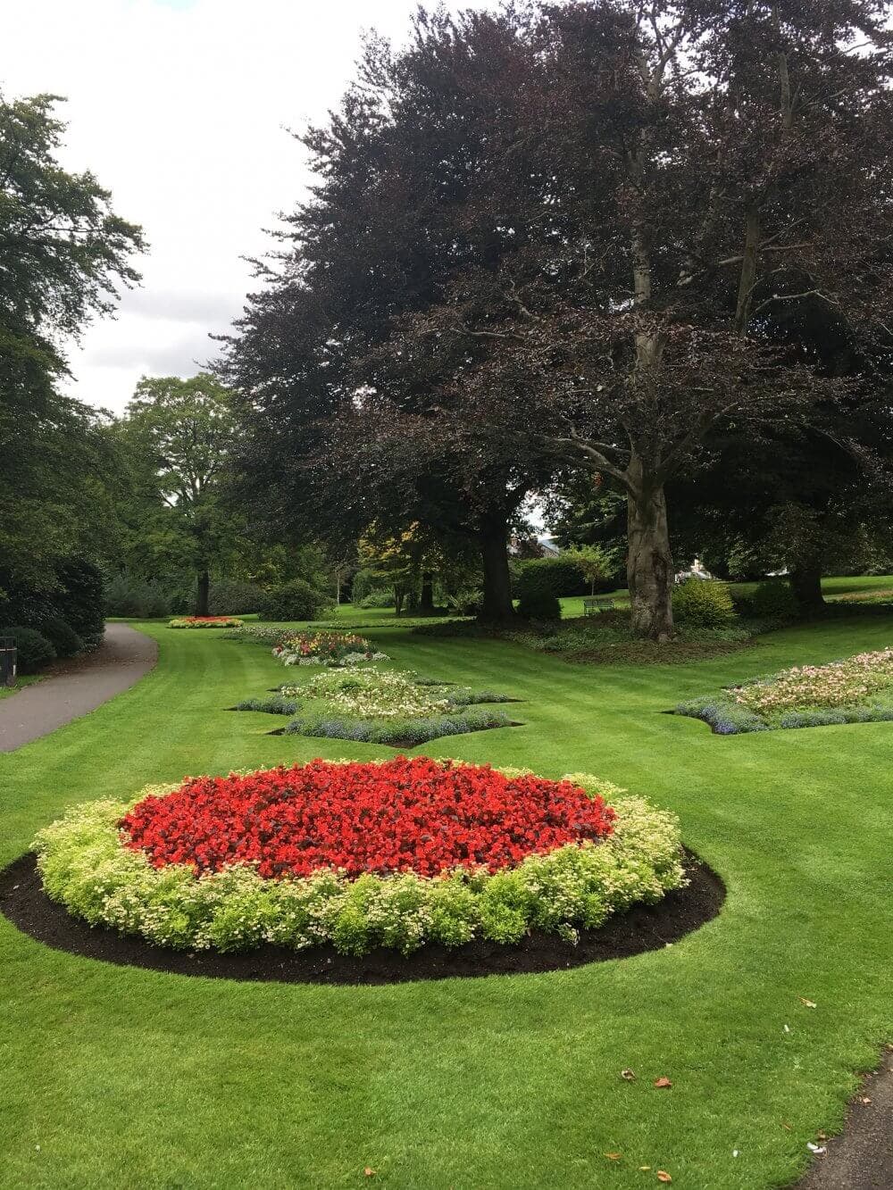 A garden with red and yellow flower beds, green lawns, and large trees under a cloudy sky. - Home Instead