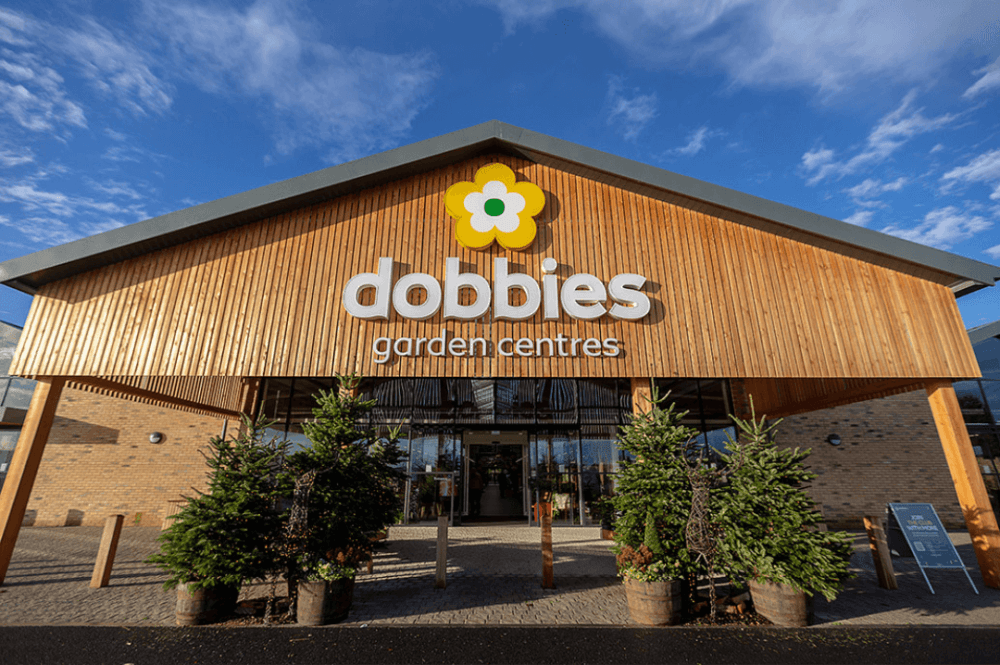 Front entrance of Dobbies Garden Centres, featuring wooden facade and surrounding green plants, under a clear blue sky. - Home Instead