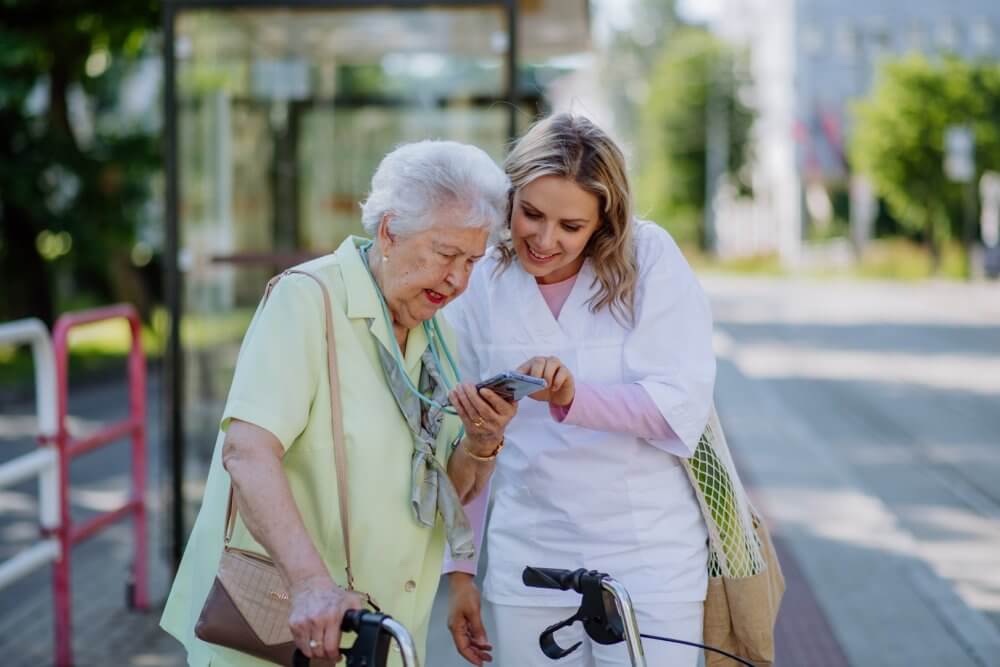 Elderly woman with walker and young woman looking at a smartphone together, smiling, on a sunny day. - Home Instead