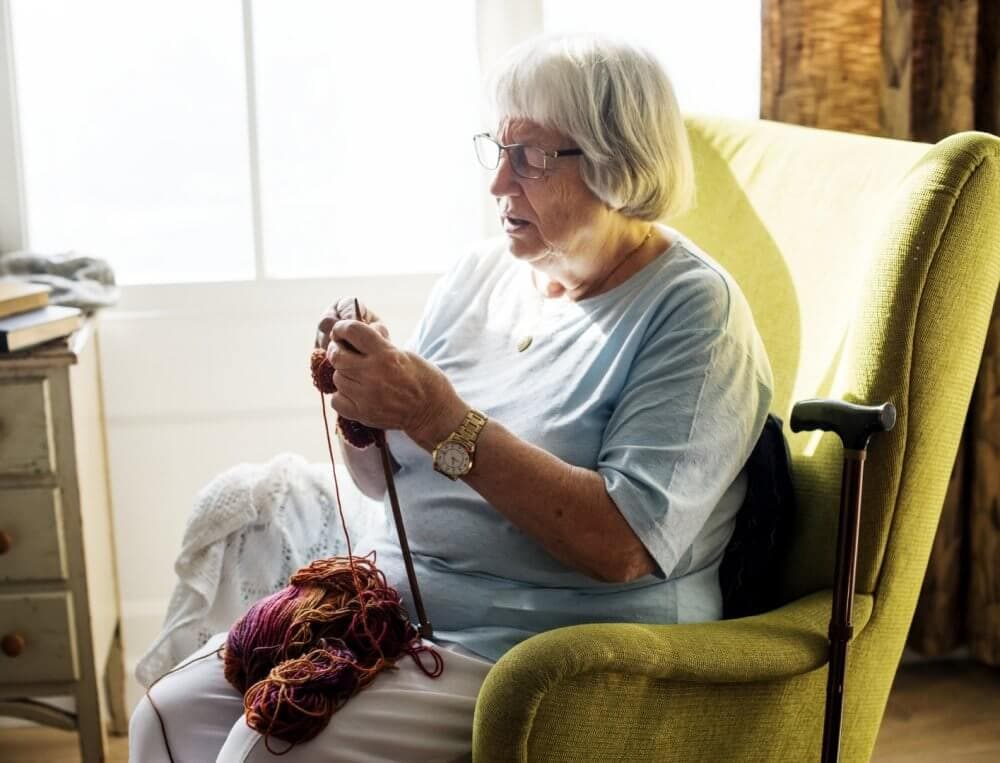 An elderly woman in a light blue shirt sits in a green armchair, knitting with colorful yarn. - Home Instead