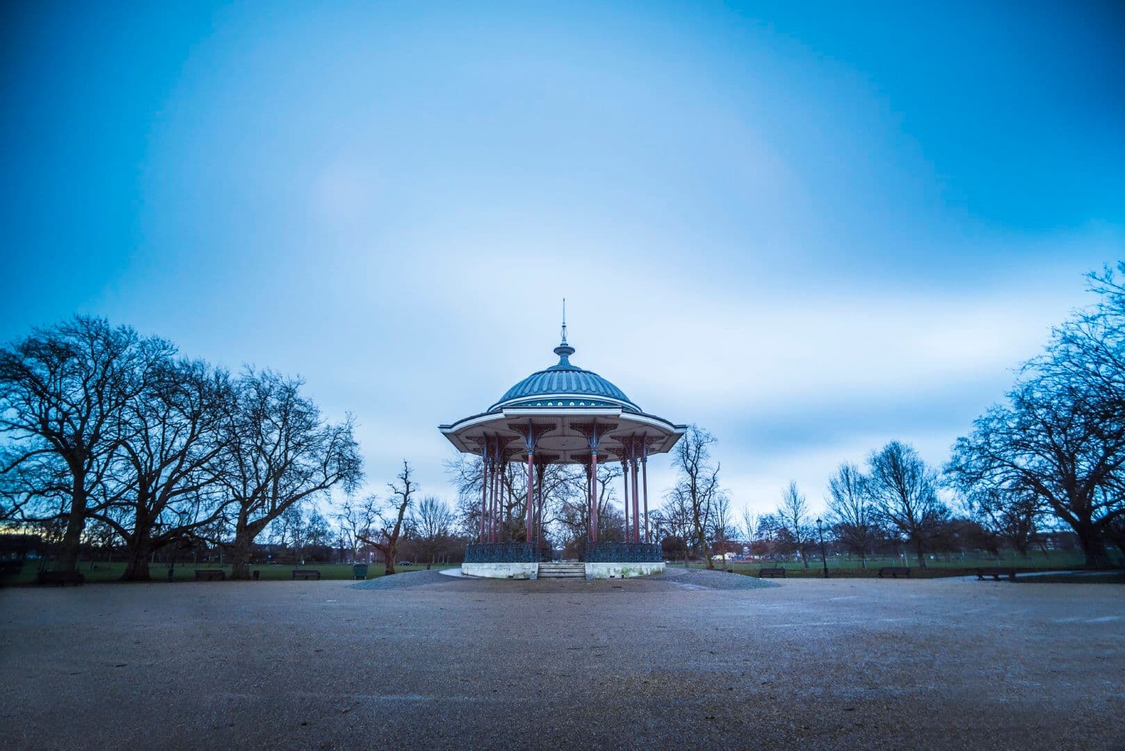 A gazebo stands in a park surrounded by bare trees under a cloudy, blue-tinged sky. - Home Instead