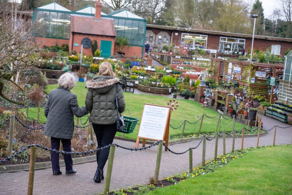 Two women walking towards a garden center, surrounded by plants and gardening supplies on a cloudy day. - Home Instead