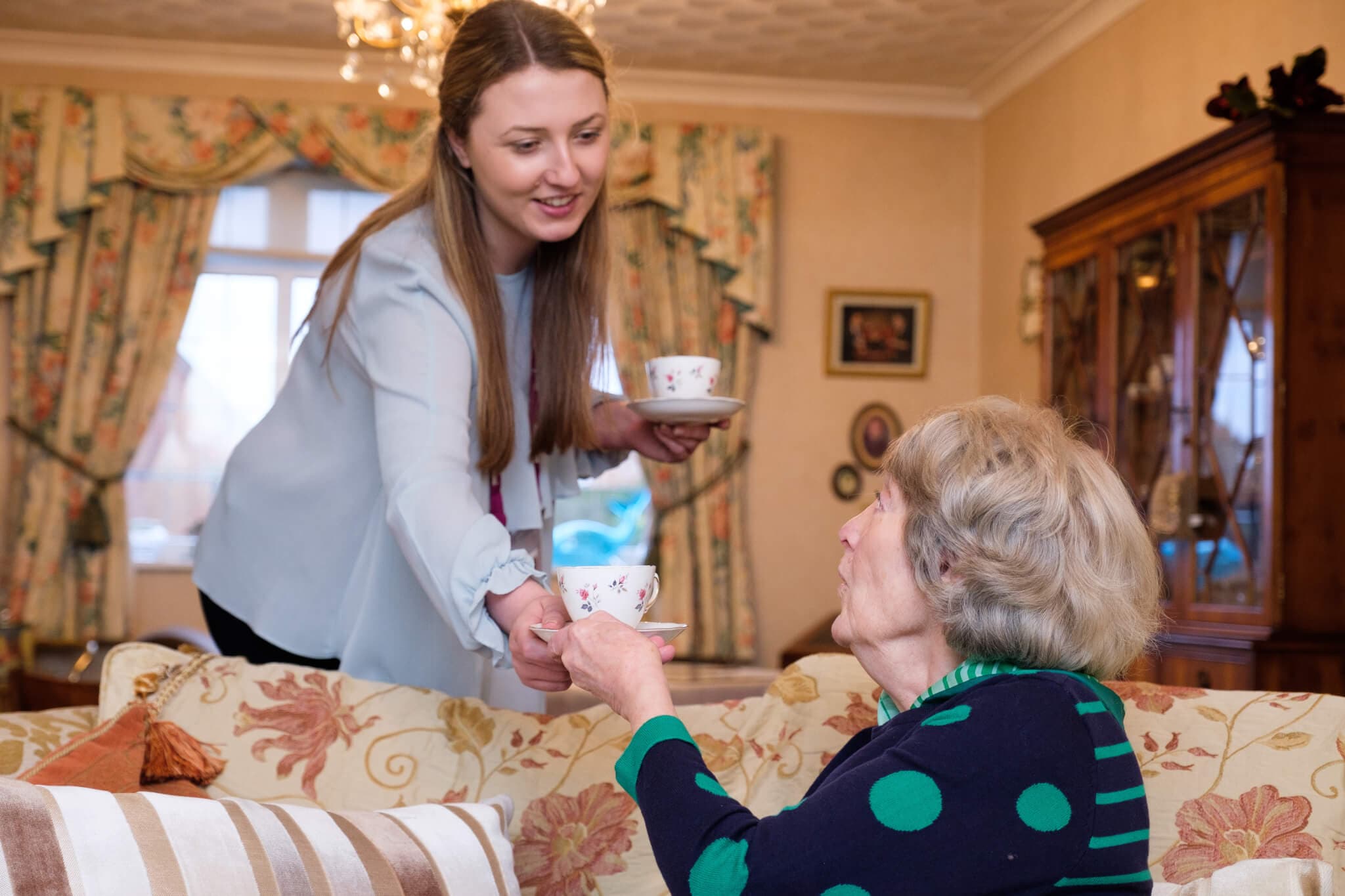 Young woman hands a teacup to an elderly woman sitting on a patterned sofa in a cozy, well-decorated living room. - Home Instead
