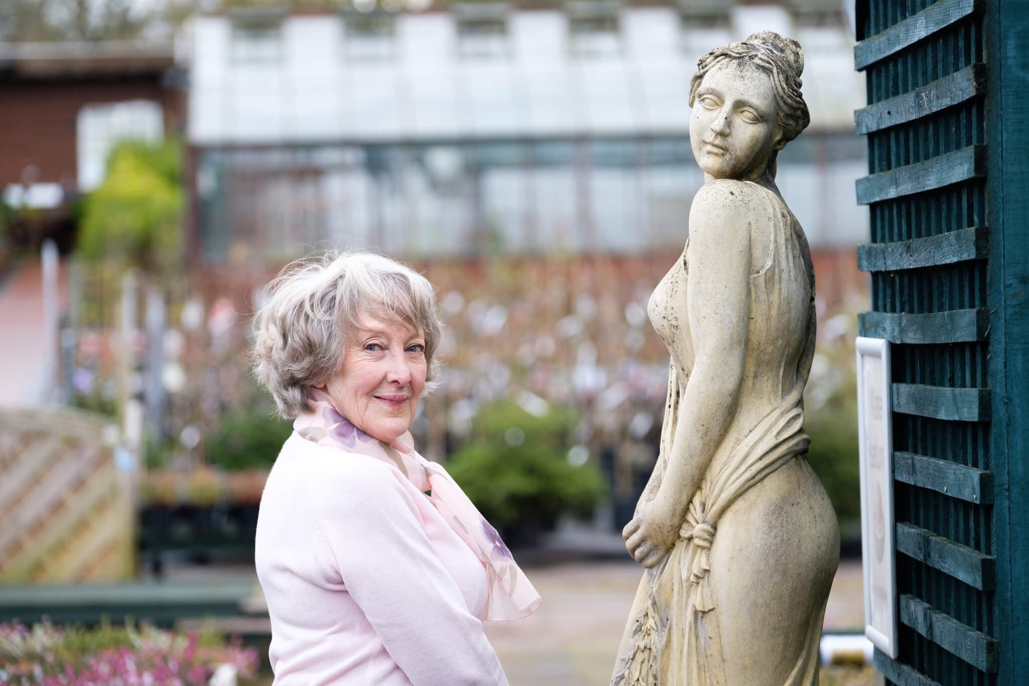 Elderly woman in pink scarf smiles next to a stone statue of a woman outside, with a greenhouse in the background. - Home Instead