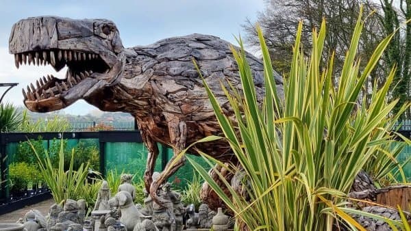A large wooden dinosaur sculpture surrounded by various plants and small stone statues in an outdoor setting. - Home Instead