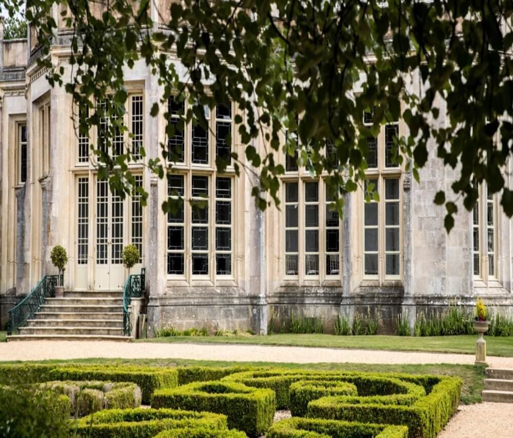 A historic stone building with large windows, a manicured garden maze, and leafy branches in the foreground. - Home Instead