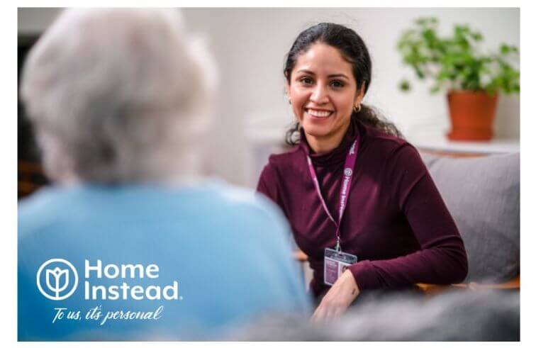 A caregiver smiles while talking to an elderly person. Logo text: "Home Instead, To us, it's personal. - Home Instead