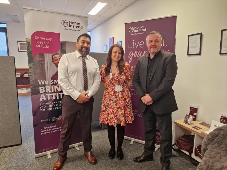 Three people standing and smiling in an office setting; two men in suits and a woman in a floral dress. - Home Instead