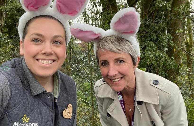 Two smiling people wearing bunny ears posing for a photo outdoors in front of greenery and a chain-link fence. - Home Instead