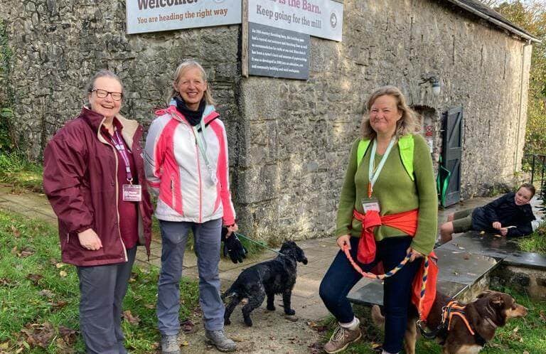 Three women with dogs stand by a stone building with a welcome sign, while one person sits nearby. - Home Instead