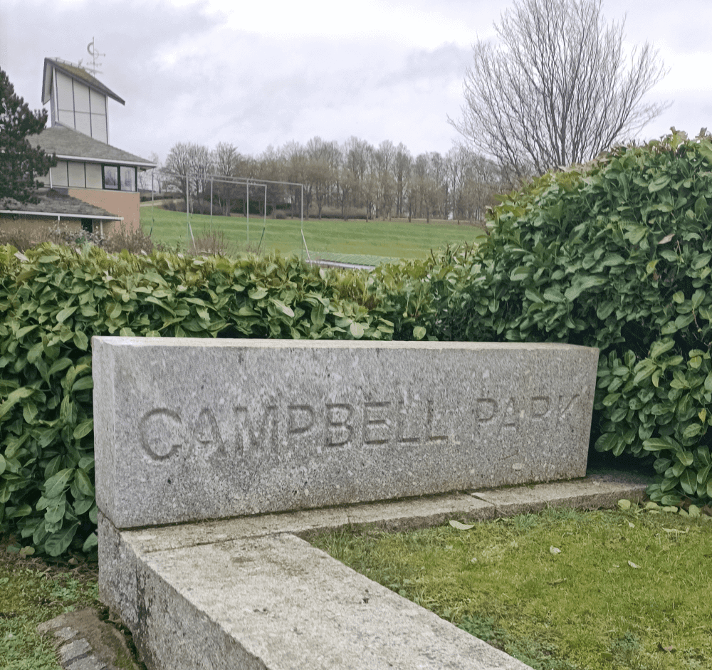 Stone sign reading "Campbell Park" with green bushes and a sports field in the background. - Home Instead
