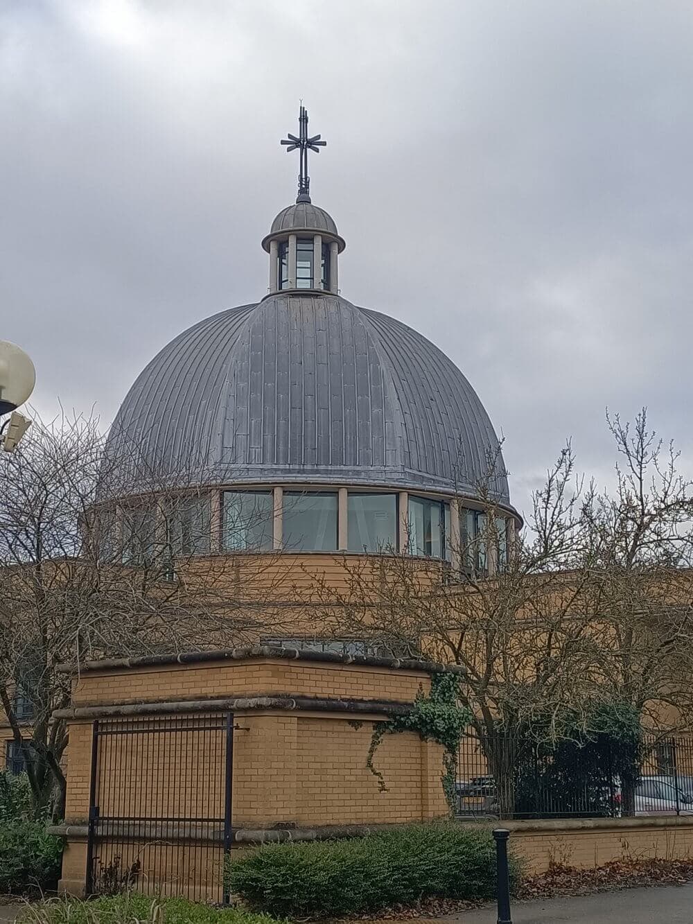 A large dome with a cross on top, surrounded by bare trees and a brick building under a cloudy sky. - Home Instead
