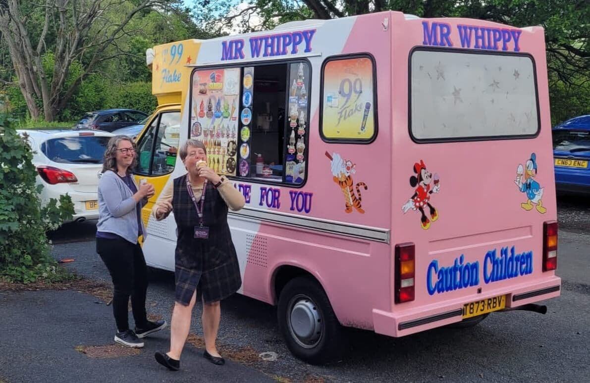 Two women enjoy ice creams from a colorful "Mr. Whippy" ice cream truck parked in a lot with trees in the background. - Home Instead