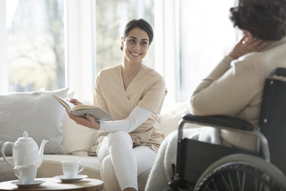 Healthcare worker holding a book and smiling at a person in a wheelchair, with tea set on the table in a bright room. - Home Instead