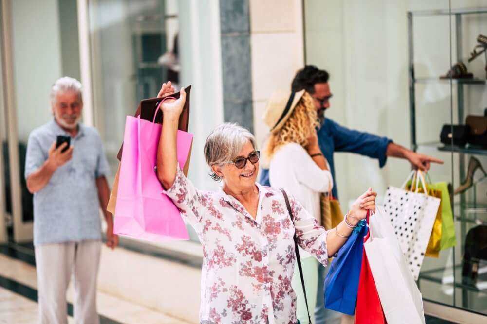 An older woman happily holding shopping bags outside a store; two people and a man on phone in the background. - Home Instead