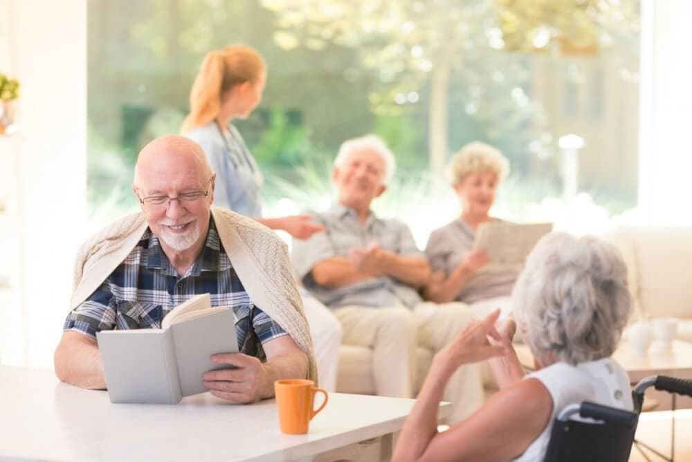 Elderly man reading a book at a table, while other seniors chat and a nurse assists in the background. - Home Instead