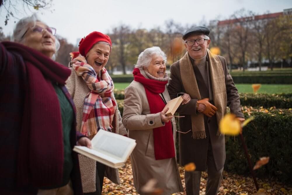 Four elderly people, dressed warmly in coats and scarves, enjoy a park while holding books as autumn leaves fall around them. - Home Instead