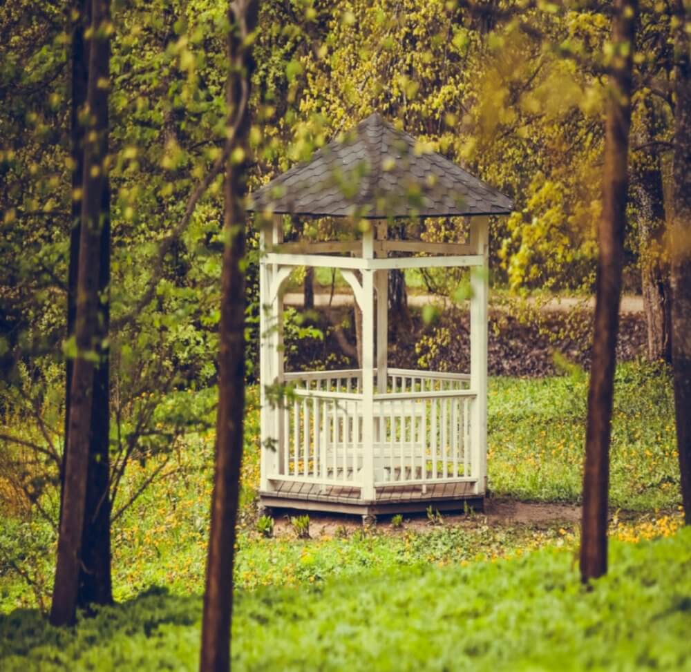 A white gazebo with a shingled roof stands amidst a lush, green forest with trees and foliage surrounding it. - Home Instead