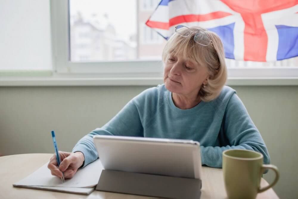 Older woman writing on paper and looking at tablet with the United Kingdom flag in the background. - Home Instead