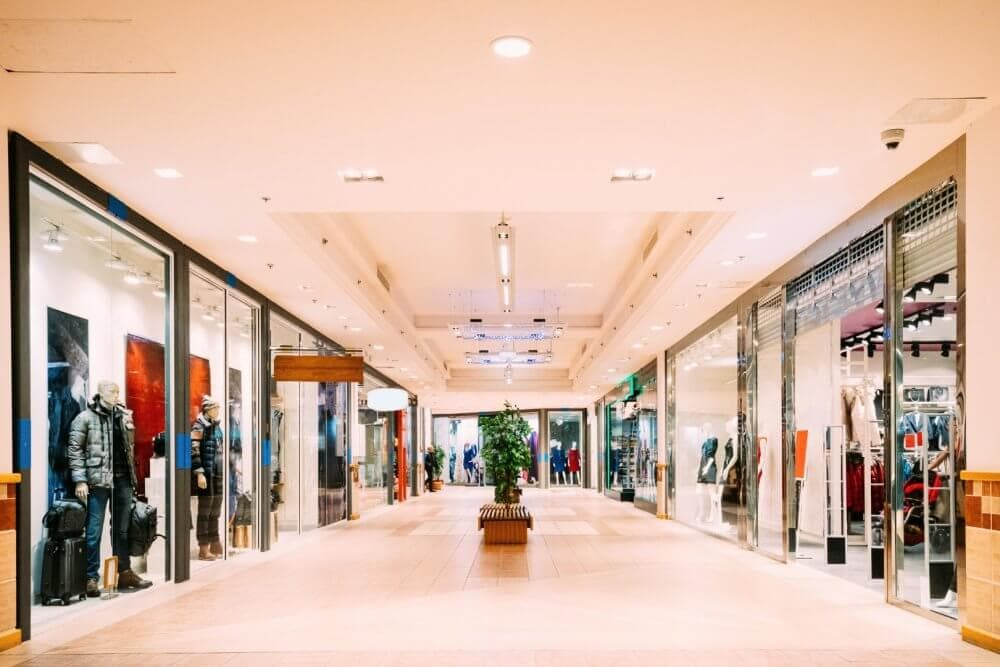 A brightly lit shopping mall corridor with stores on both sides, mannequins in display windows, and a small tree in the center. - Home Instead
