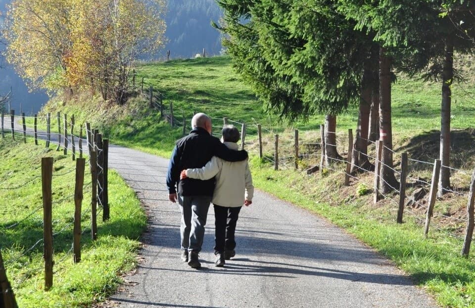 Elderly couple walking arm in arm on a paved path in the countryside, surrounded by trees and greenery. - Home Instead