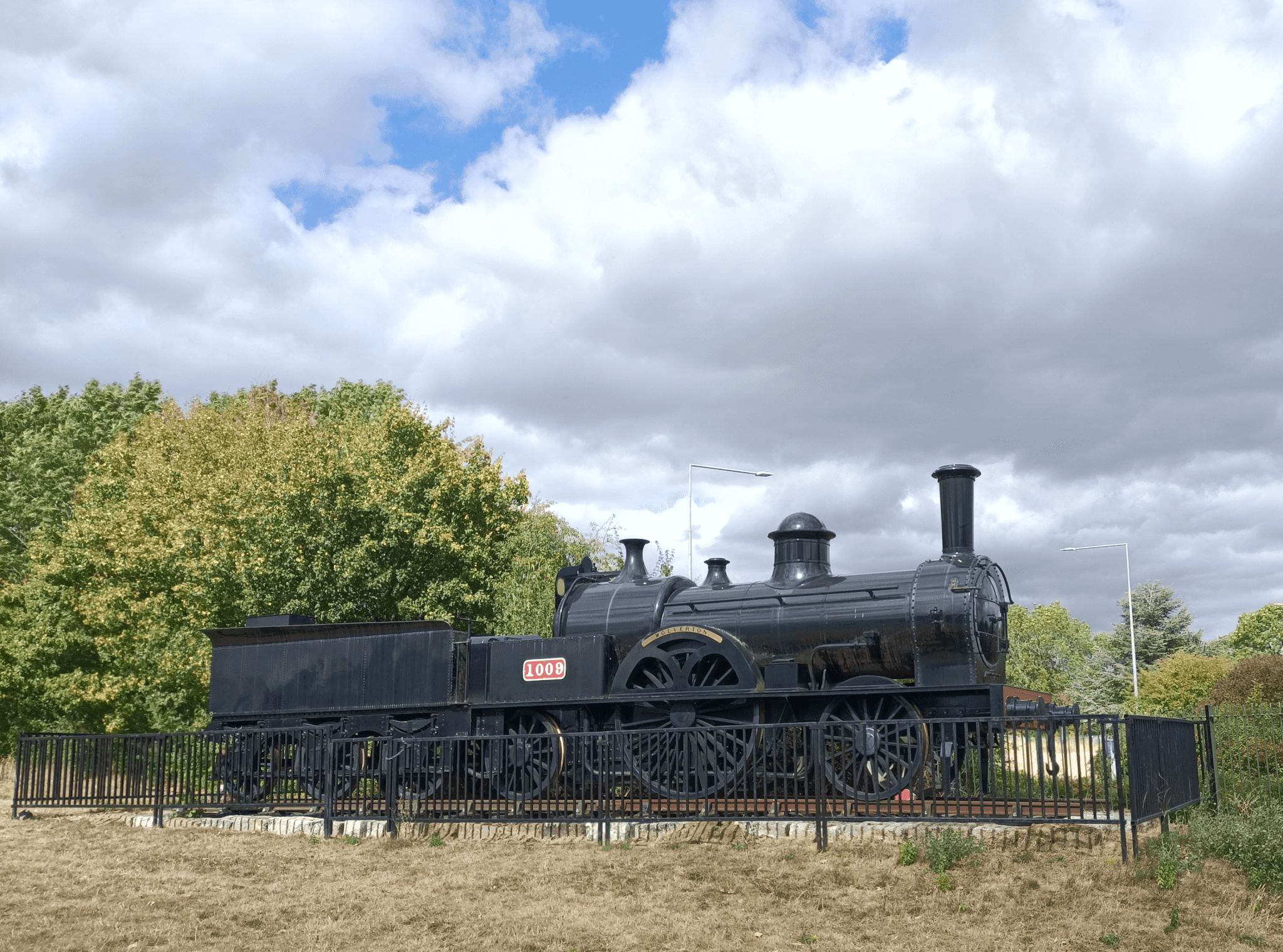A black vintage steam locomotive on display, surrounded by a black fence and greenery under a cloudy sky. - Home Instead