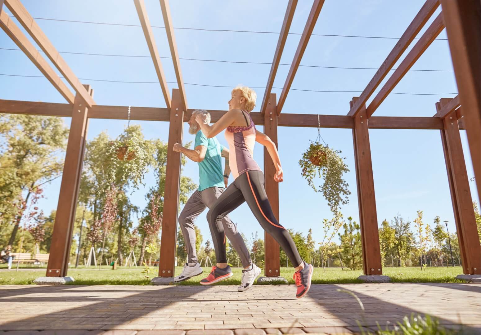 Two people jogging under a wooden pergola in a sunny park, dressed in athletic wear. Trees and hanging plants in the background. - Home Instead