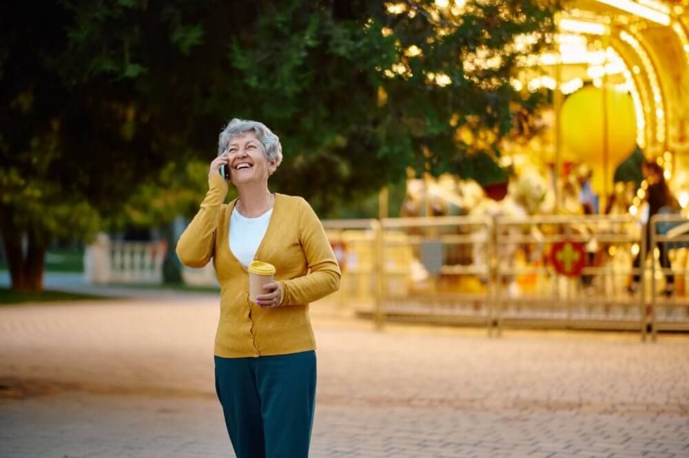 An elderly woman in a yellow sweater laughs while on the phone, holding a coffee cup near a brightly lit carousel. - Home Instead