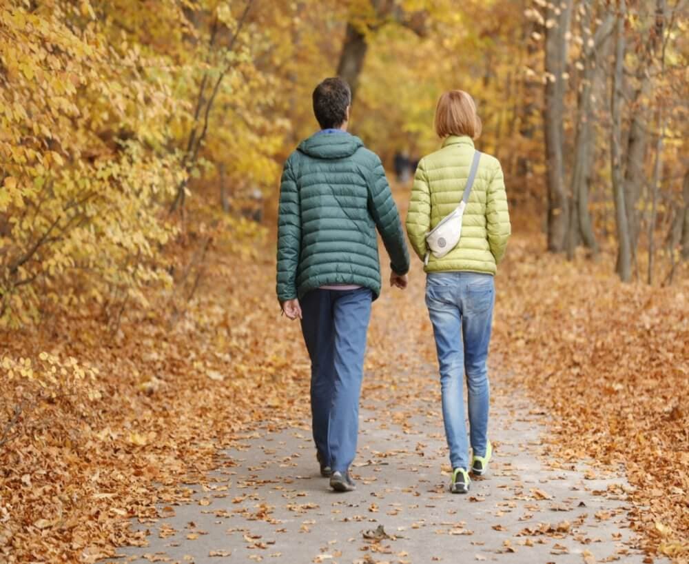 A couple in jackets walks hand-in-hand along a leaf-covered path through an autumn forest. - Home Instead