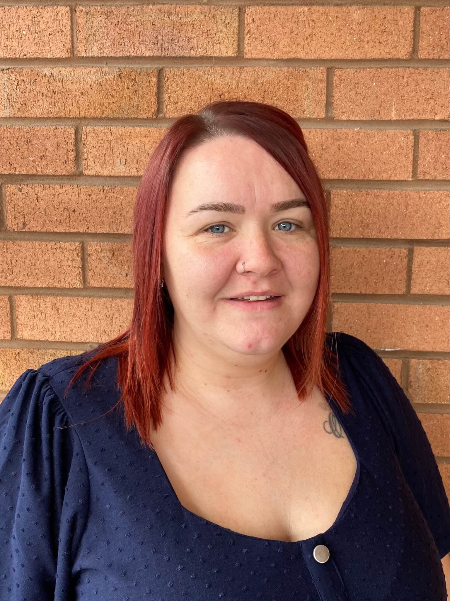 A woman with red hair and a navy blue top stands in front of a brick wall, smiling at the camera. - Home Instead