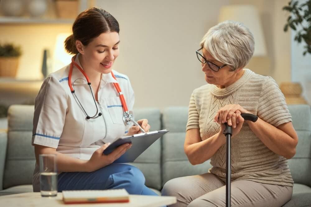 A nurse holding a clipboard speaks to an elderly woman using a cane, both seated in a cozy, well-lit room. - Home Instead Bournemouth & Christchurch