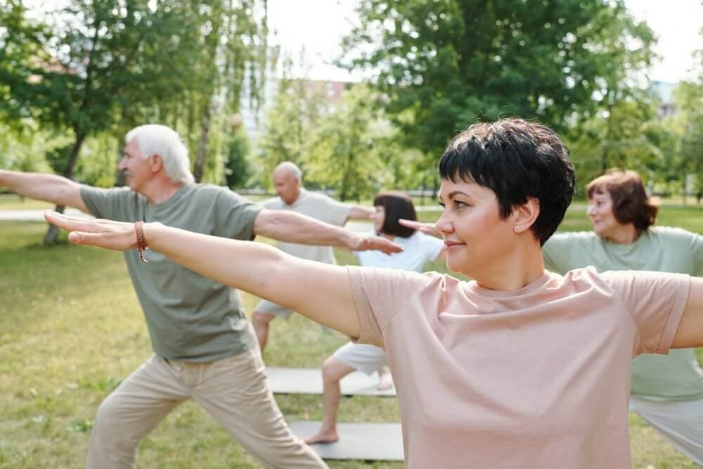 A group of people practicing yoga in a park, performing the Warrior II pose on yoga mats. - Home Instead