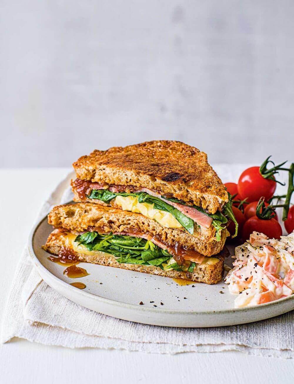 A grilled sandwich with greens, egg, tomatoes, and sauce, served with coleslaw and cherry tomatoes on a beige plate. - Home Instead