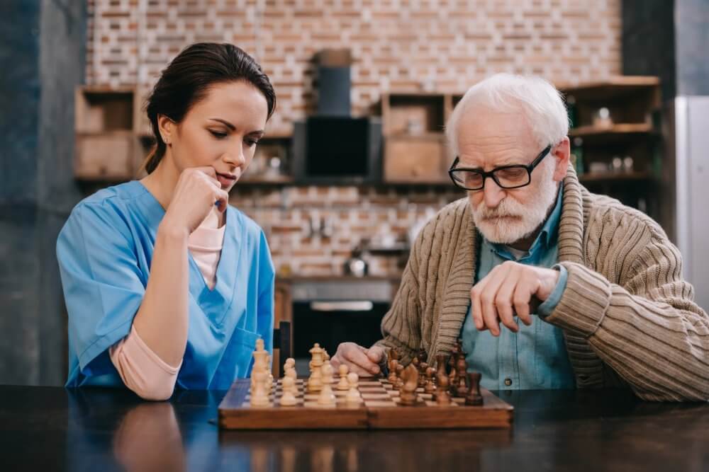 An elderly man and a nurse play a game of chess in a cozy room with a brick wall in the background. - Home Instead Bournemouth & Christchurch
