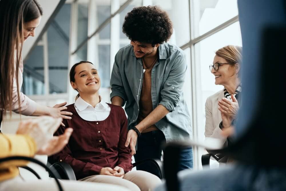 A group of people joyfully gathers around a seated woman, offering support and congratulating her. - Home Instead Bournemouth & Christchurch