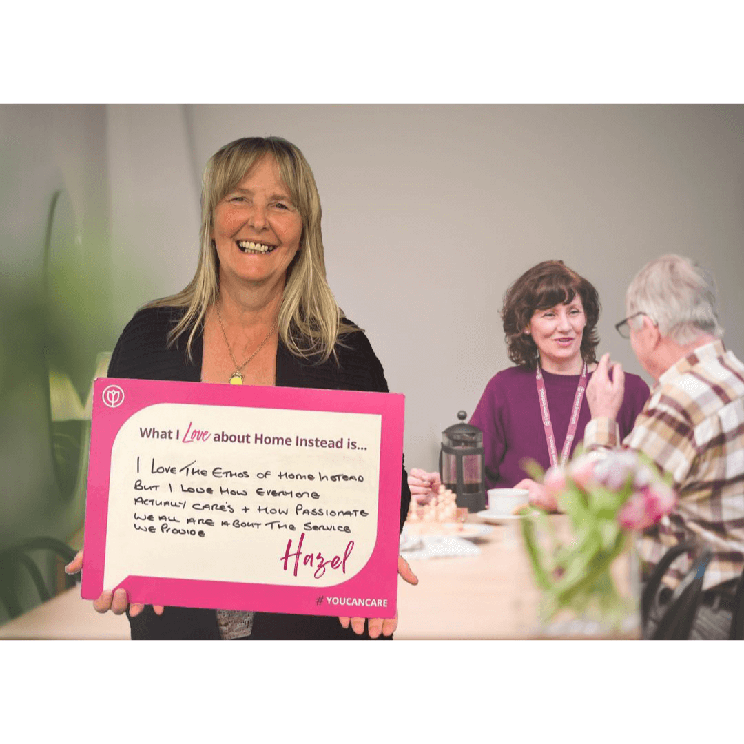 A woman smiling and holding a sign while two people converse in the background at a table. - Home Instead