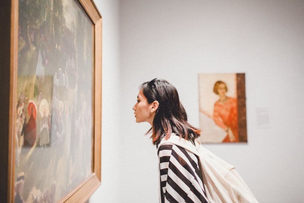 Person with a backpack leaning in to closely examine a painting in an art gallery. Another painting is in the background. - Home Instead Bournemouth & Christchurch