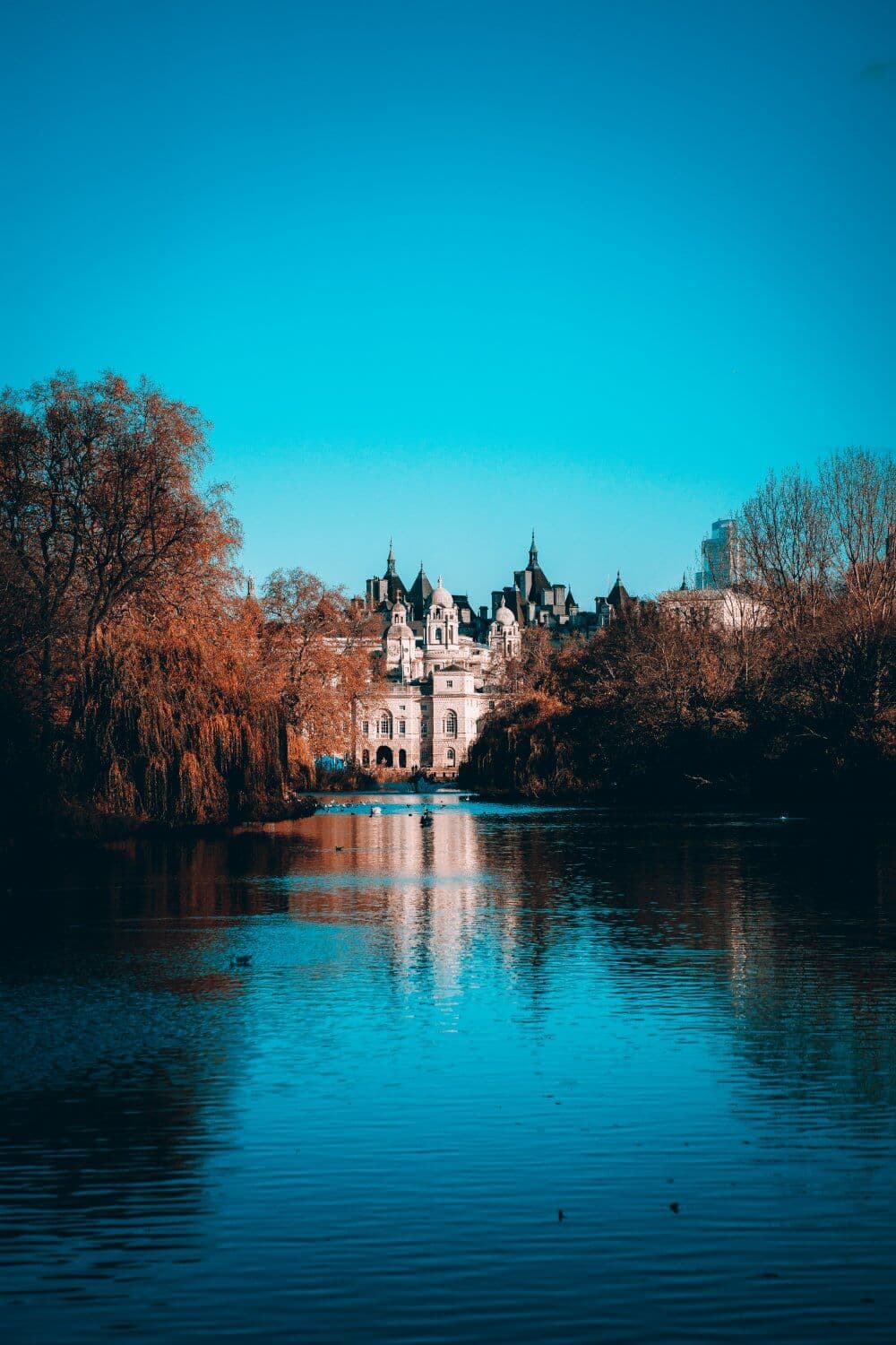 Scenic view of a castle-like building in the distance with reflections on a calm lake surrounded by autumn trees. - Home Instead