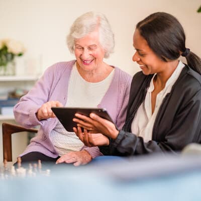 An elderly woman and a younger woman smiling and looking at a tablet together, sitting in a cozy room. - Home Instead